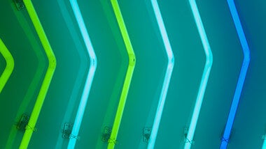 Image dans les tons bleus. Ce sont des néons en forme de flèches indiquant la droite. Ils sont accrochés à un mur qui reflète leur lumière. Les néons sont verts ou bleus.