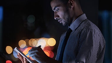 Un homme de profil, en chemise et cravate, qui écrit sur une tablette avec un stylet. Fond sombre avec des points de lumière flous.
