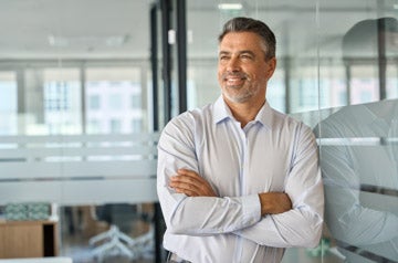Happy mid aged older professional business man, mature latin executive manager, 50 years old investor standing in office arms crossed leaning at glass wall looking away thinking of success concept.