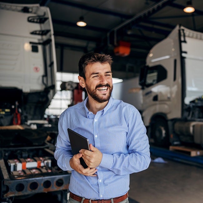 Smiling chief standing in auto park and holding tablet. In background are trucks. Firm for import and export.