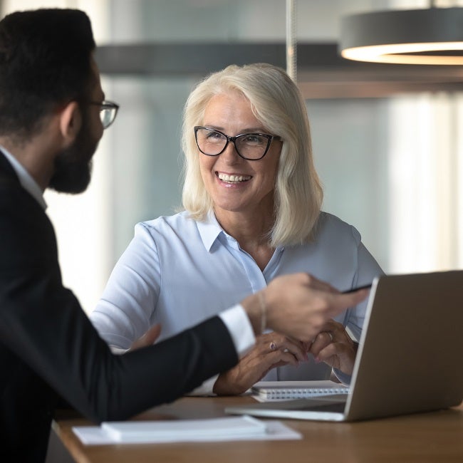 Businessman consulting mature businesswoman client about loan, insurance or investment, using laptop, pointing at screen, young man helping senior colleague with software, employee presenting project