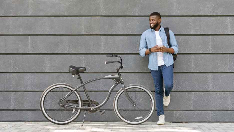 Handsome afro guy with backgpack and smartphone standing near his bike, leaning on gray urban wall, copy space